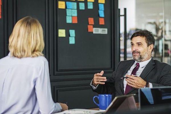 older business man speaking with a blond haired woman with sticky notes in background