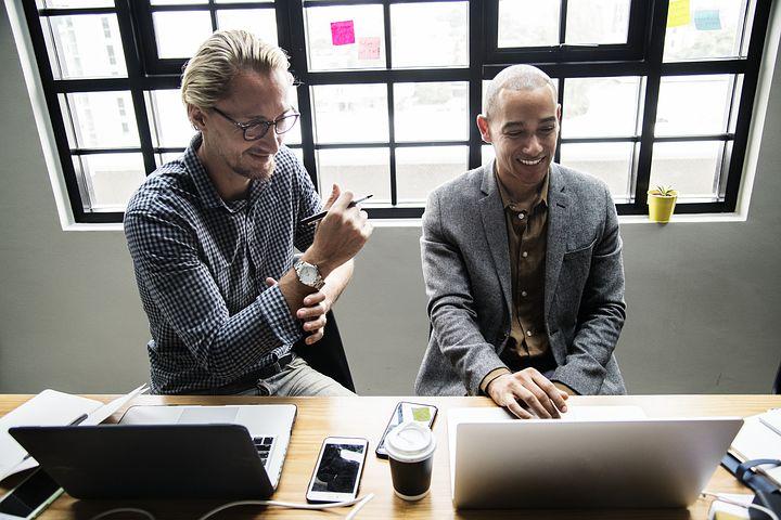 two male workers sitting next to each other in front of their laptops smiling