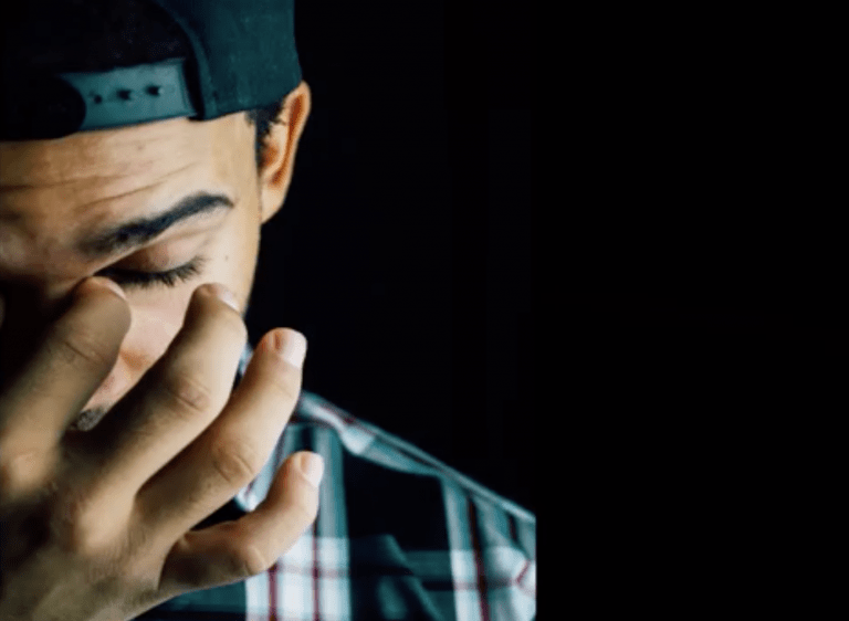 man with hat turned backwards with hand pinching his nose as if he has a headache or stress