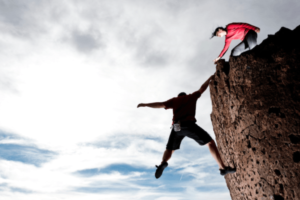 man almost falling off of cliff but holding on with one hand with other man leaning over